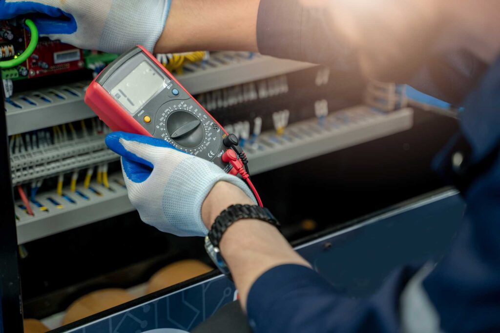 A person wearing gloves uses a digital multimeter to test electrical connections inside an open control panel.