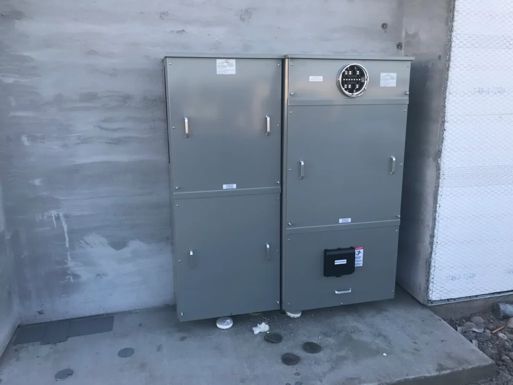 Two gray electrical utility boxes are mounted against a concrete wall; one features a round meter on its upper right door.