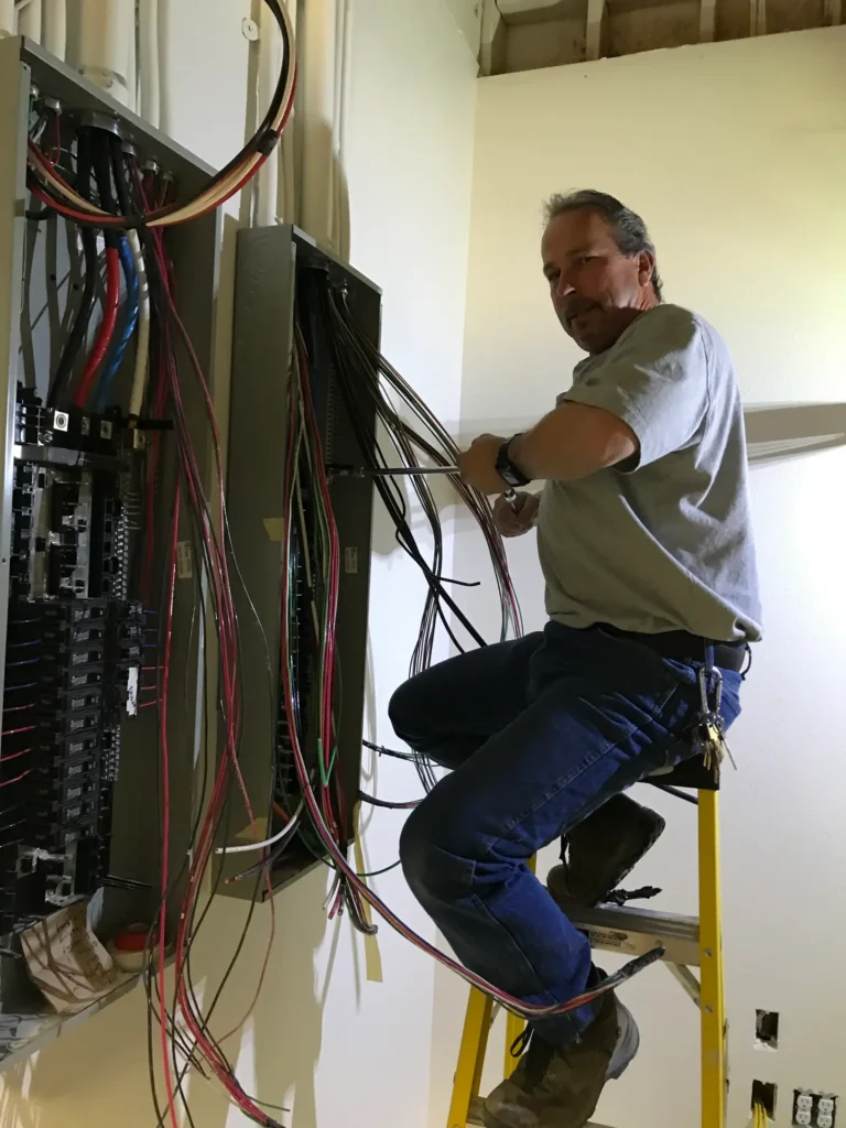 An electrician stands on a ladder and works on wiring inside an electrical panel mounted on the wall.