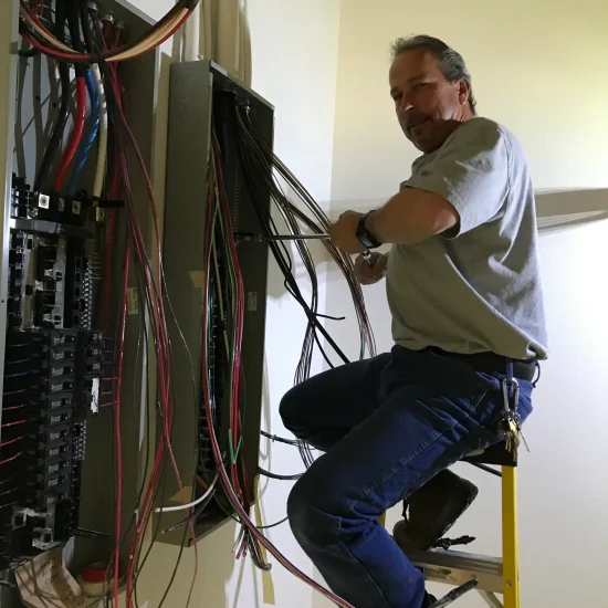Goodkin (8) An electrician stands on a ladder and works on wiring inside an electrical panel mounted on the wall.