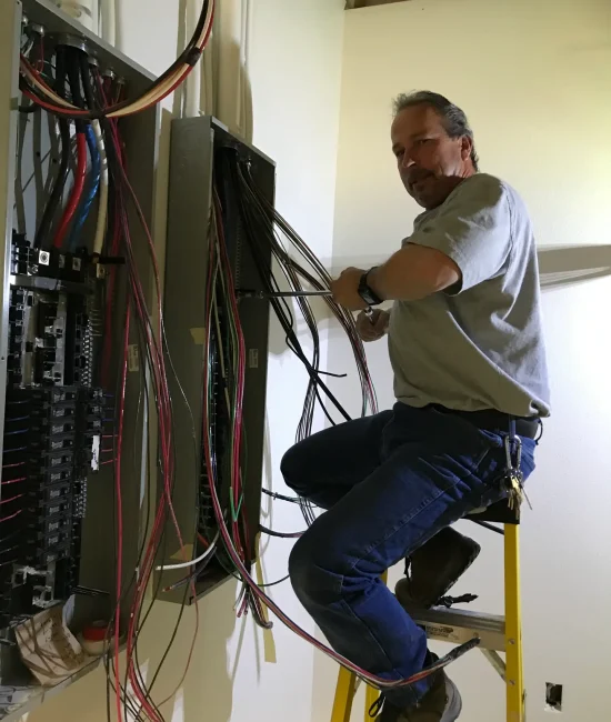 Goodkin (8) An electrician stands on a ladder and works on wiring inside an electrical panel mounted on the wall.