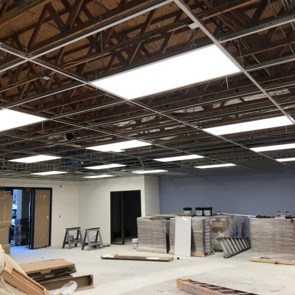 Office under renovation with exposed ceiling, LED panel lights installed, construction materials, and equipment scattered on the floor.
