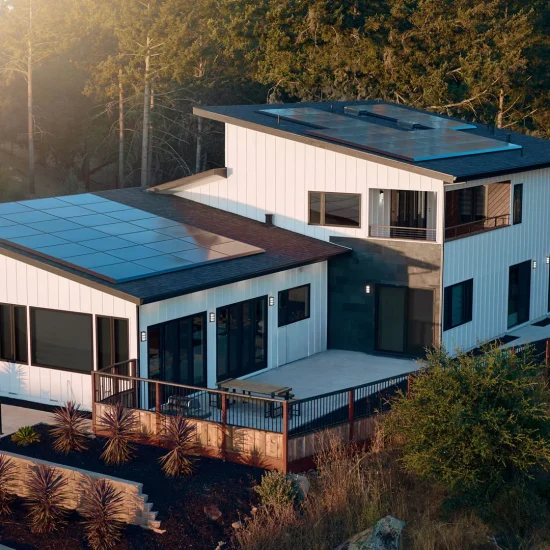 Modern two-story house with solar panels on the roof from Tesla Energy, large windows, a red car parked nearby, and a landscaped garden in a wooded area.