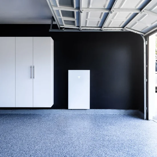 A clean garage interior features white storage cabinets, a Tesla Powerwall battery from Tesla Energy mounted on a black wall, and a car parked outside in the driveway—showcasing modern energy solutions.