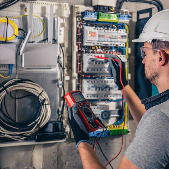 man-an-electrical-technician-working-in-a-switchb-A9E28TE.jpg An electrician wearing a helmet and gloves uses a multimeter to test an electrical panel with organized wires and equipment on a wall.