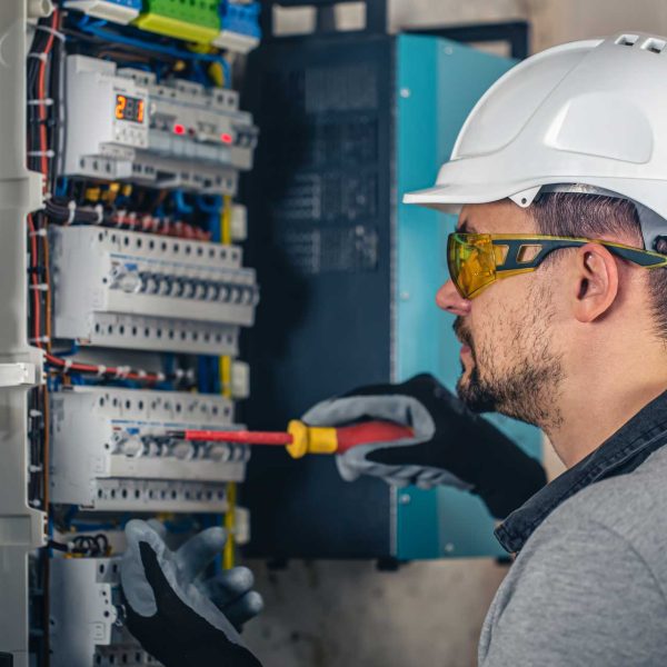 man-an-electrical-technician-working-in-a-switchb-UJH642G.jpg A man wearing a hard hat and safety glasses uses a screwdriver to work on an electrical panel with multiple wires and switches.