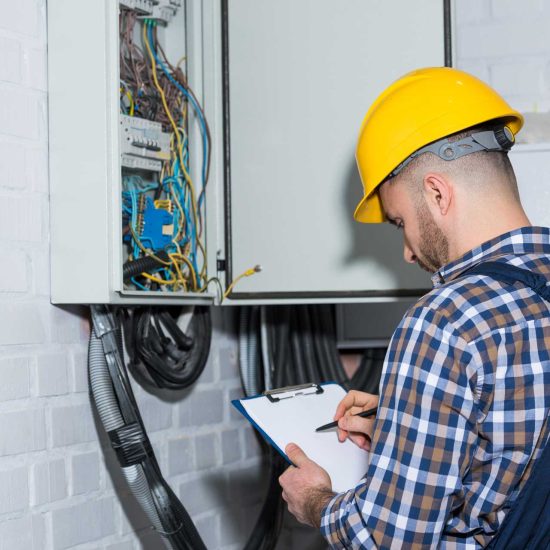 professional-electrician-inspecting-wires-in-elect-YEB3RXQ.jpg An electrician wearing a yellow hard hat and checkered shirt writes on a clipboard while inspecting an open electrical panel on a white brick wall.